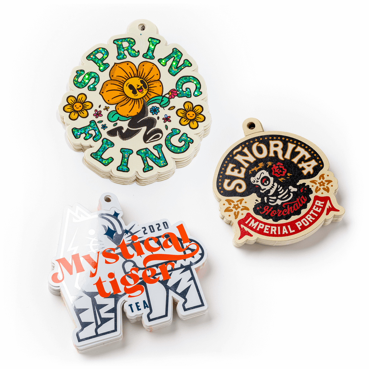 Three colorful, illustrated keychains: "Spring Fling" with flowers, "Señorita" with a skull, and "Mystical Tiger" with bold text.