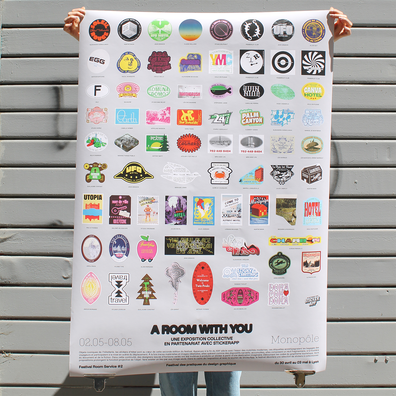 man holding a poster showing colorful designs and badges, standing in front of a garage