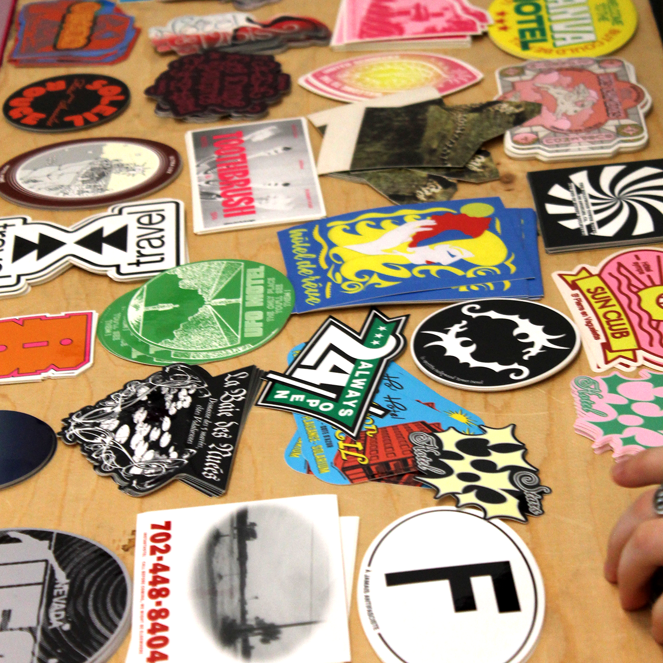 hand pointing at colorful die-cut stickers with travel themes and the text "TRAVEL" and "SUN CLUB", laying on a wooden table