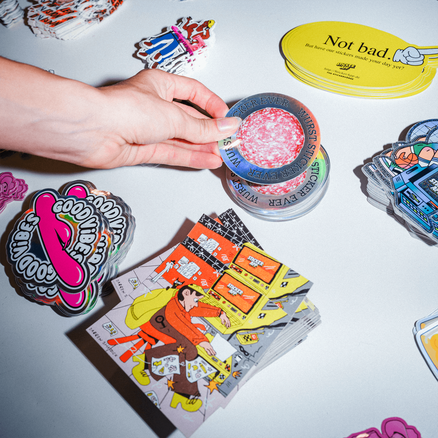 Hand holding a round holographic salami sticker above a table of die-cut stickers with different finishes