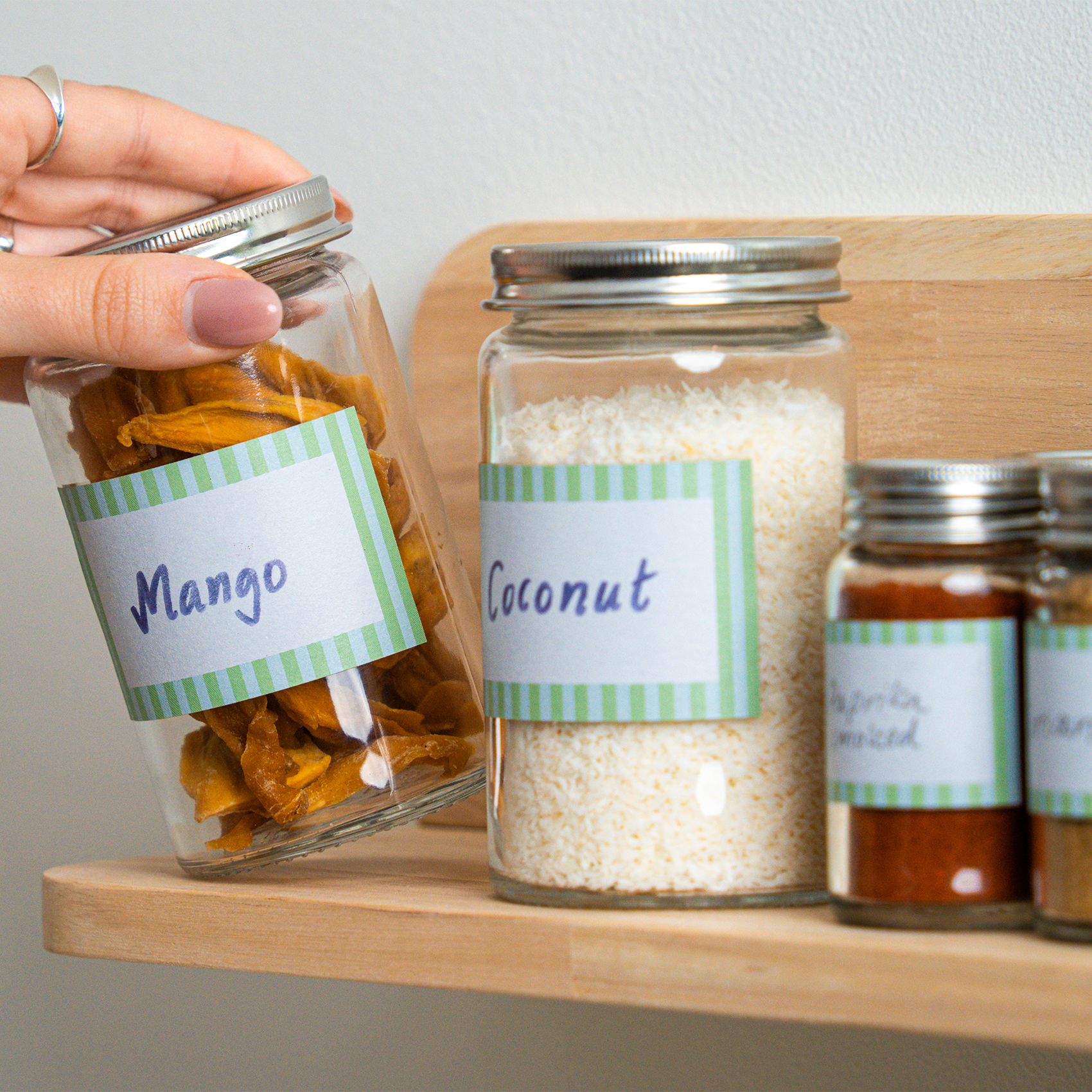 A hand placing a glass jar labeled “Mango” onto a wooden shelf next to other labeled jars containing coconut and spices.