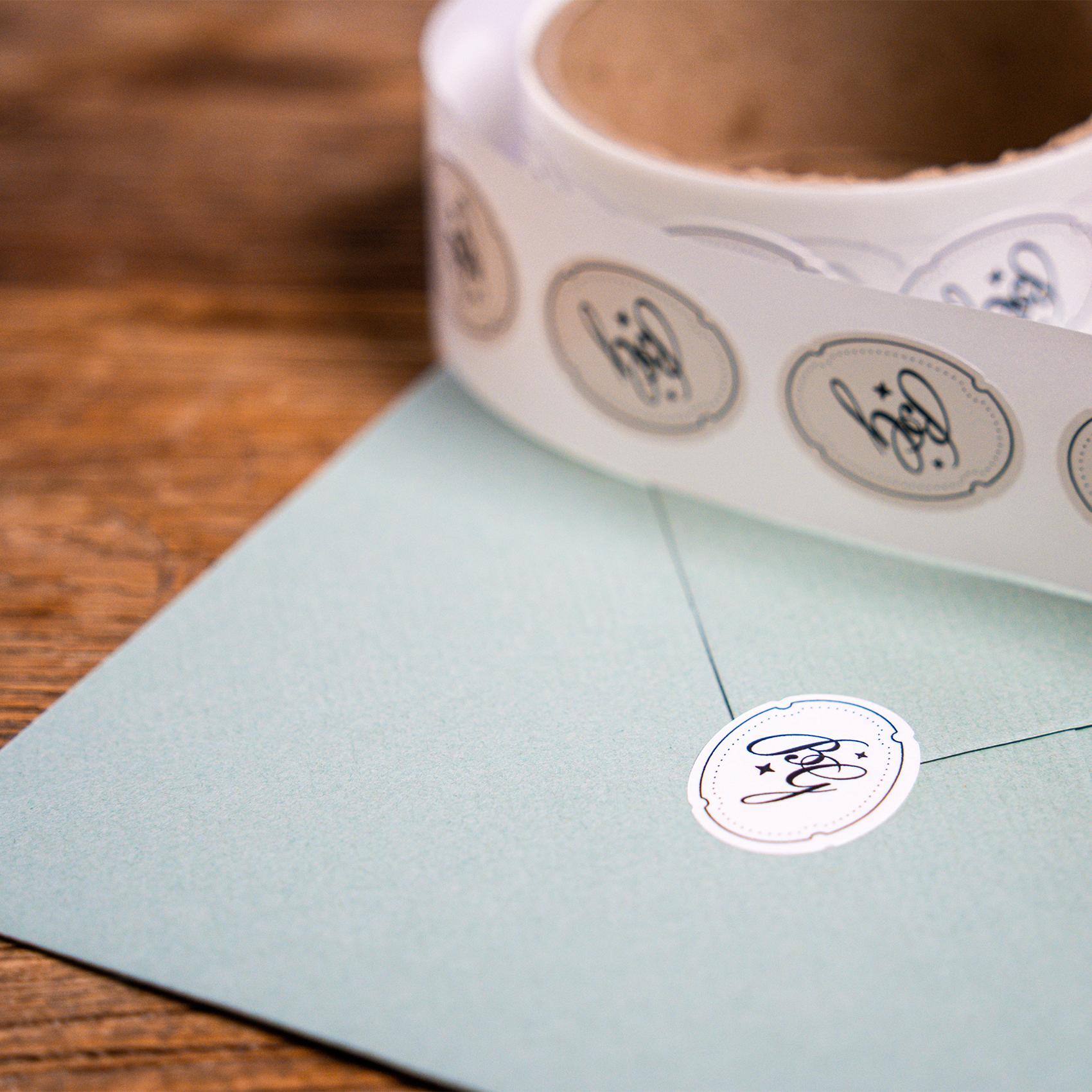 A roll of round custom stickers placed next to a light blue envelope sealed with a matching logo sticker on a wooden table.