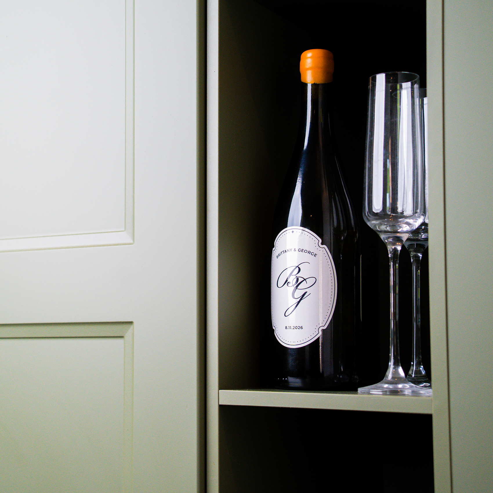 A dark wine bottle with an range cap standing inside a cabinet next to two tall champagne glasses.