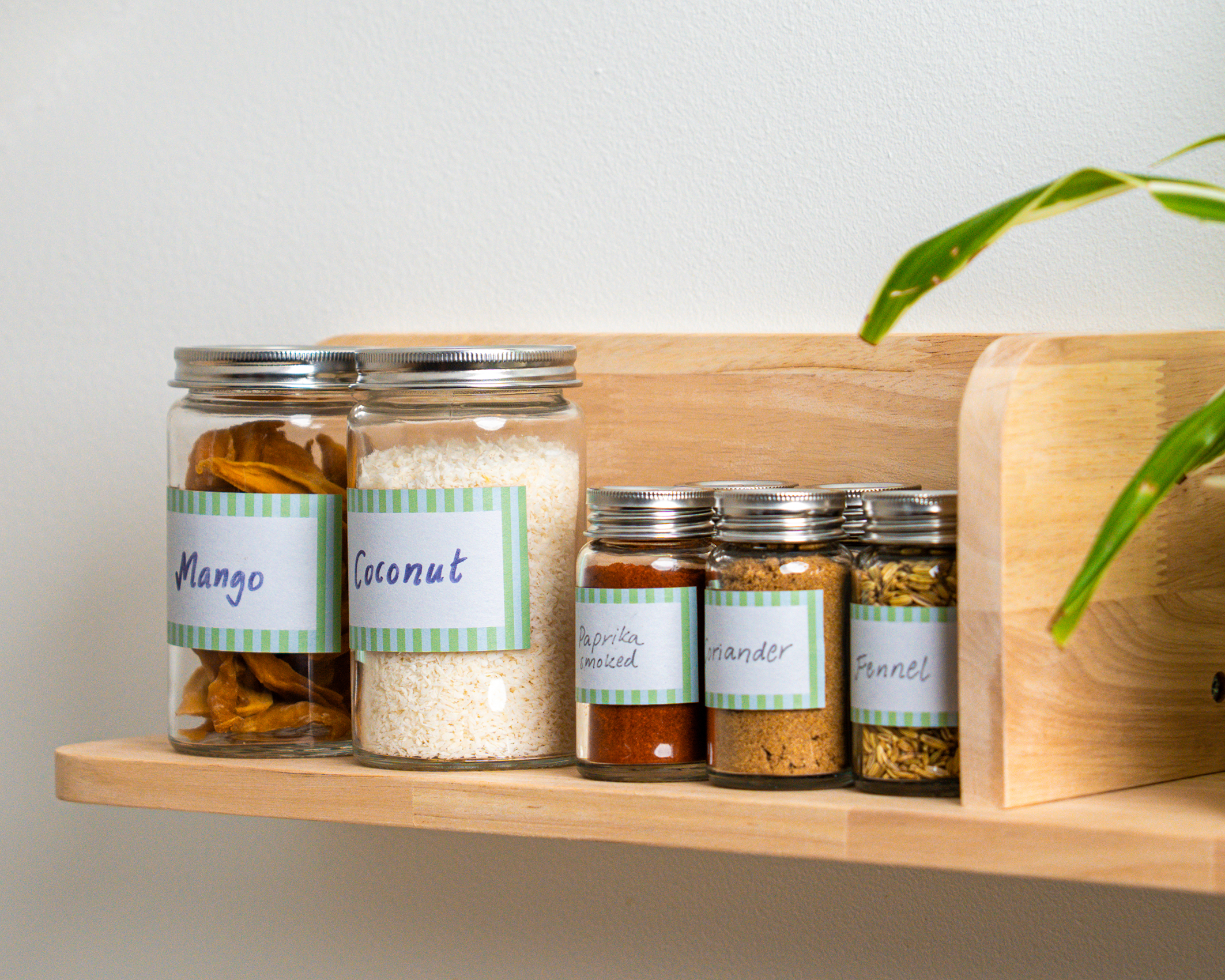 Glass jars with handwritten labels placed on a wooden kitchen shelf. The jars contain dried mango, coconut, and various spices including smoked paprika, coriander, and fennel seeds.