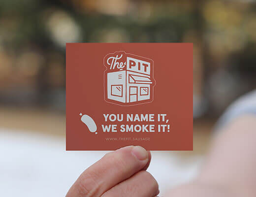 A person holding up a kiss cut sticker that says "the pit you name it, we smoke it!"